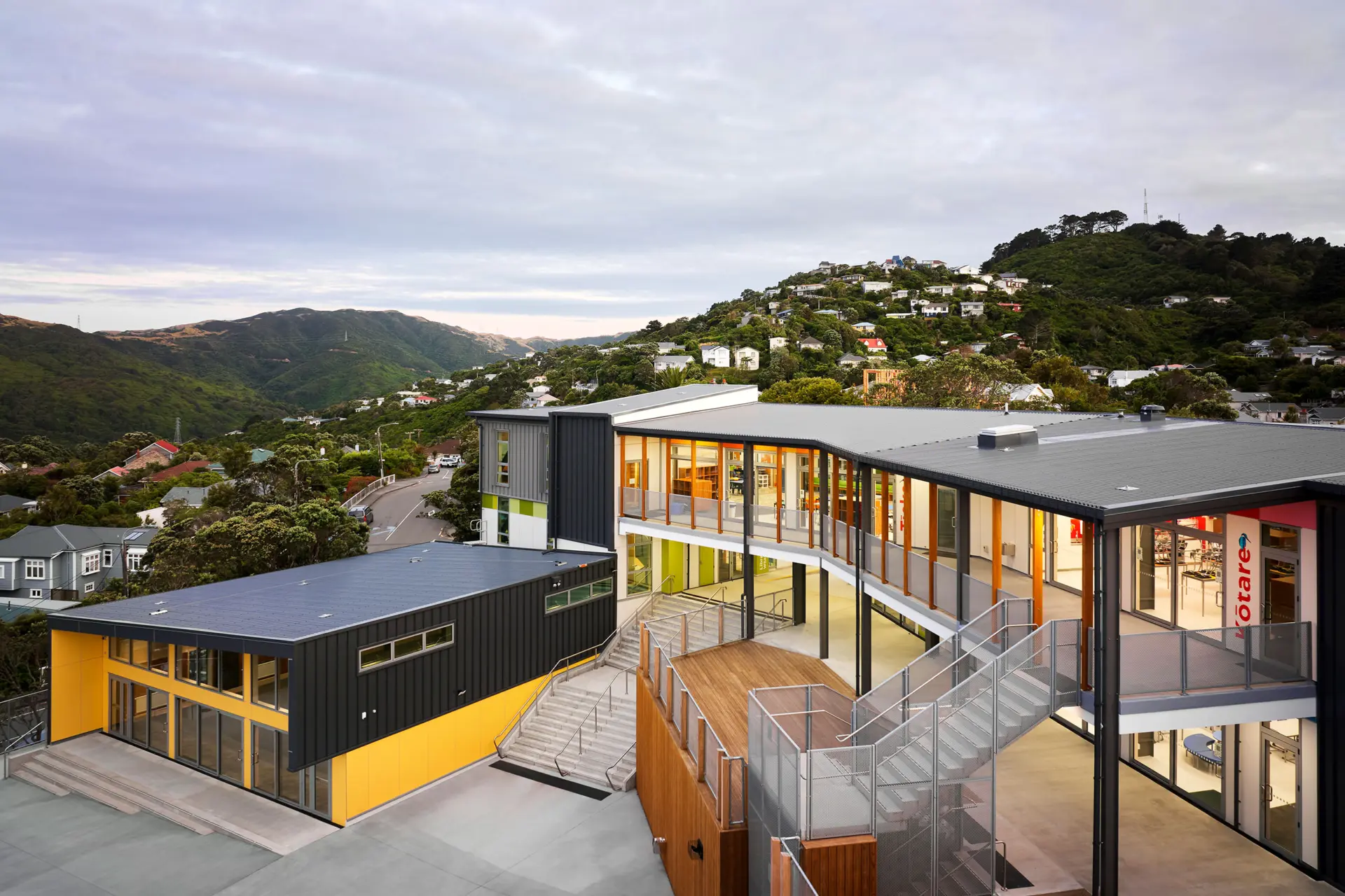A new two-storey community building with dark and yellow cladding, timber decking and full-height glazing, set against a bush-covered hillside.